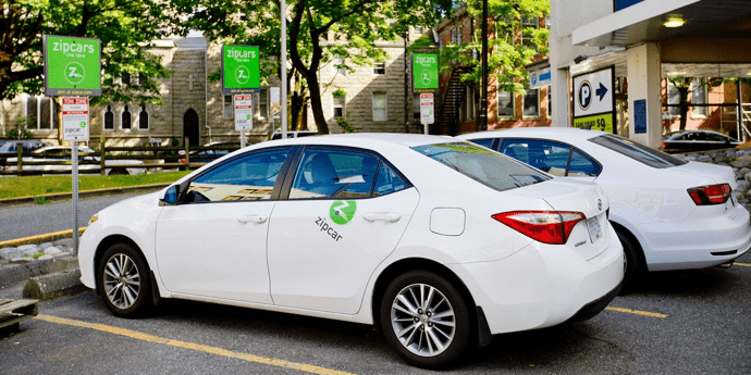 Two white compact cars parked at parking spots marked for ZipCar pick-up/drop-off.
